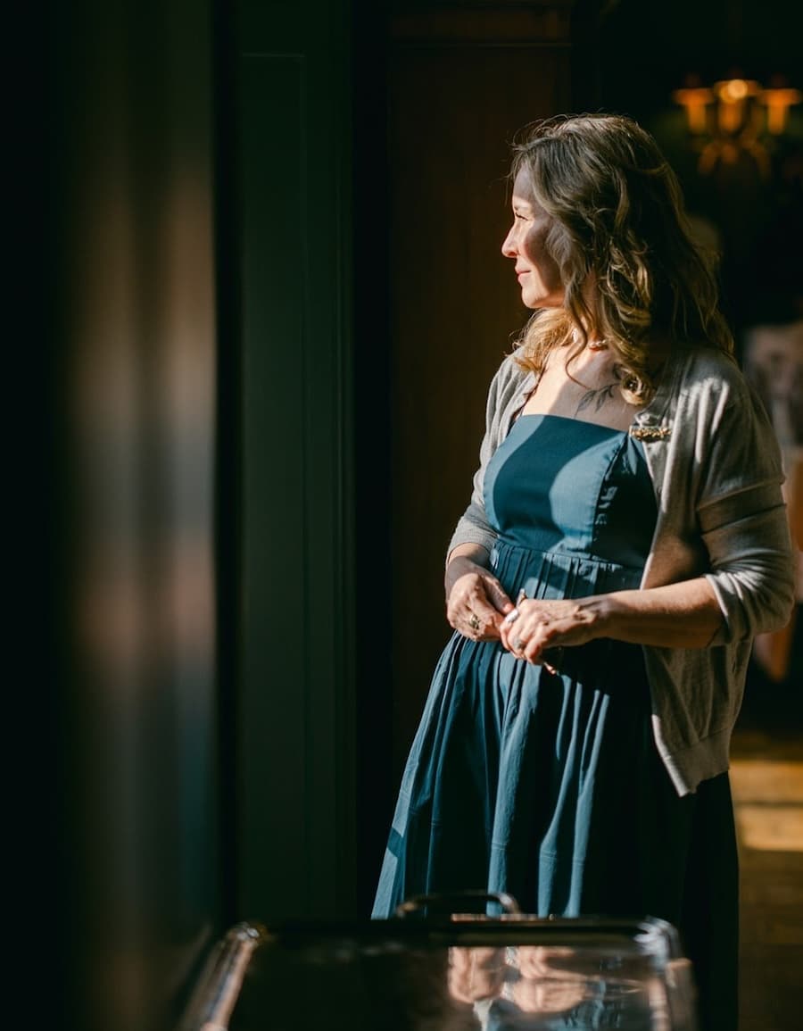 Woman in a blue dress and gray cardigan stands by a window, looking outside with sunlight on her face.