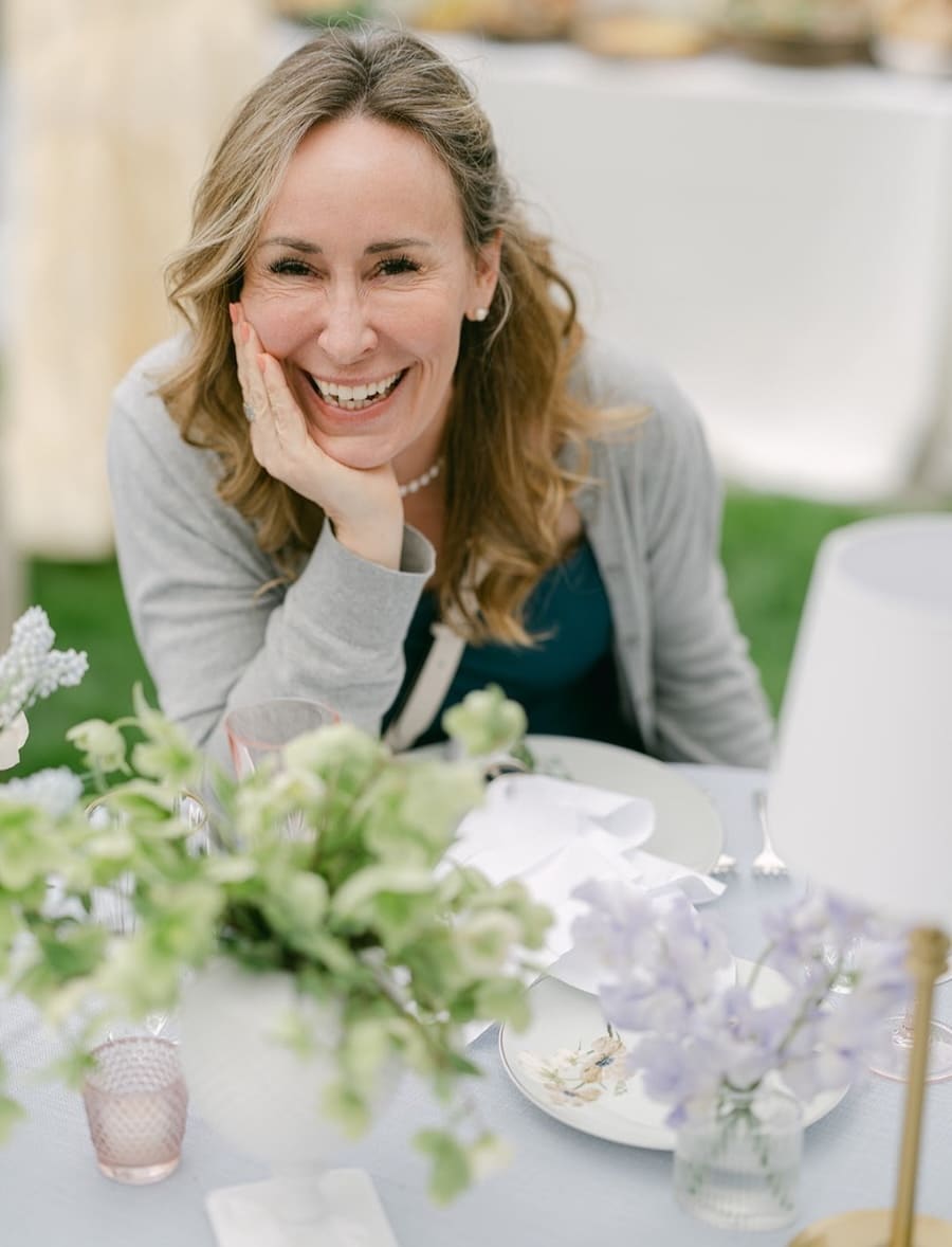 A woman smiling and sitting at a table decorated with flowers and tableware outdoors.