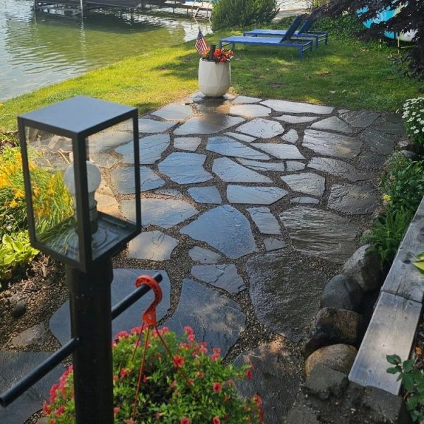 A wet stone patio by a lake, surrounded by flowers, greenery, and a black lamp post, with lounge chairs nearby.